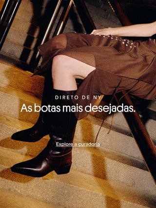 Banner com modelo sentada em escada usando botas de couro preto de cano médio e salto baixo. Sobreposição de texto: “Direto de NY. As botas mais desejadas.” e chamada “Explore a curadoria”.