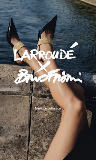 Pernas cruzadas sobre mureta à beira d'água usando scarpin slingback preto de bico fino com tornozeleira dourada – Larroudé x Bruno Frisoni